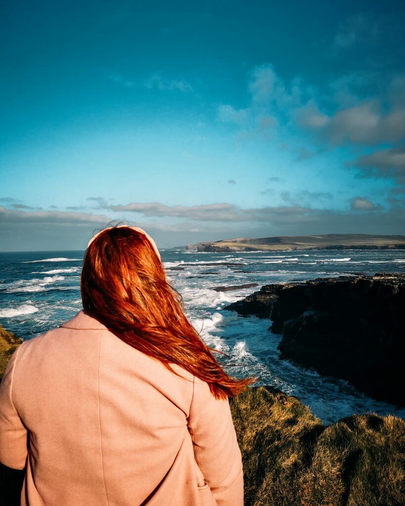 Irish woman with red hair and wears a pink coat looks out over the Irish coastline.