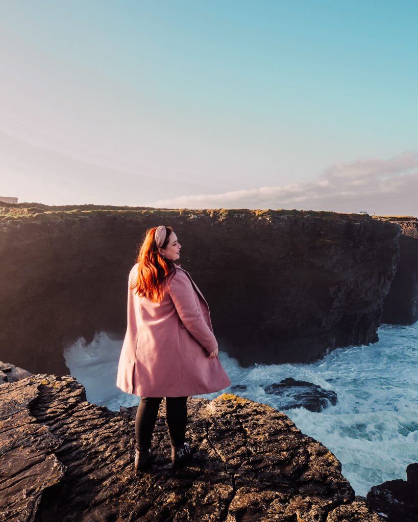 Nicola Lavin, Irish Travel Blogger, wears a pink coat and stands at the edge of Kilkee cliffs in County Clare Ireland overlooking the crashing waves of the Atlantic Ocean.