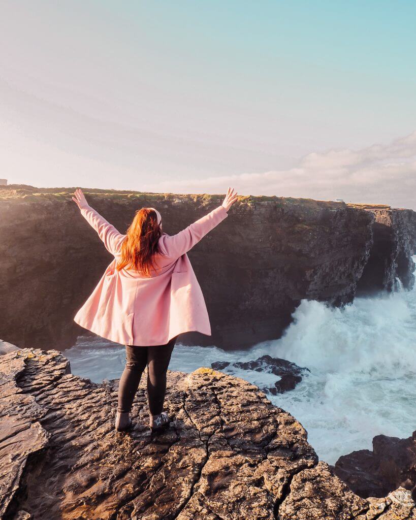 Irish Woman with red hair wearing a pink coat has her hands up in the air as she stands on a cliff edge as the waves of the Atlantic Ocean crash below her.