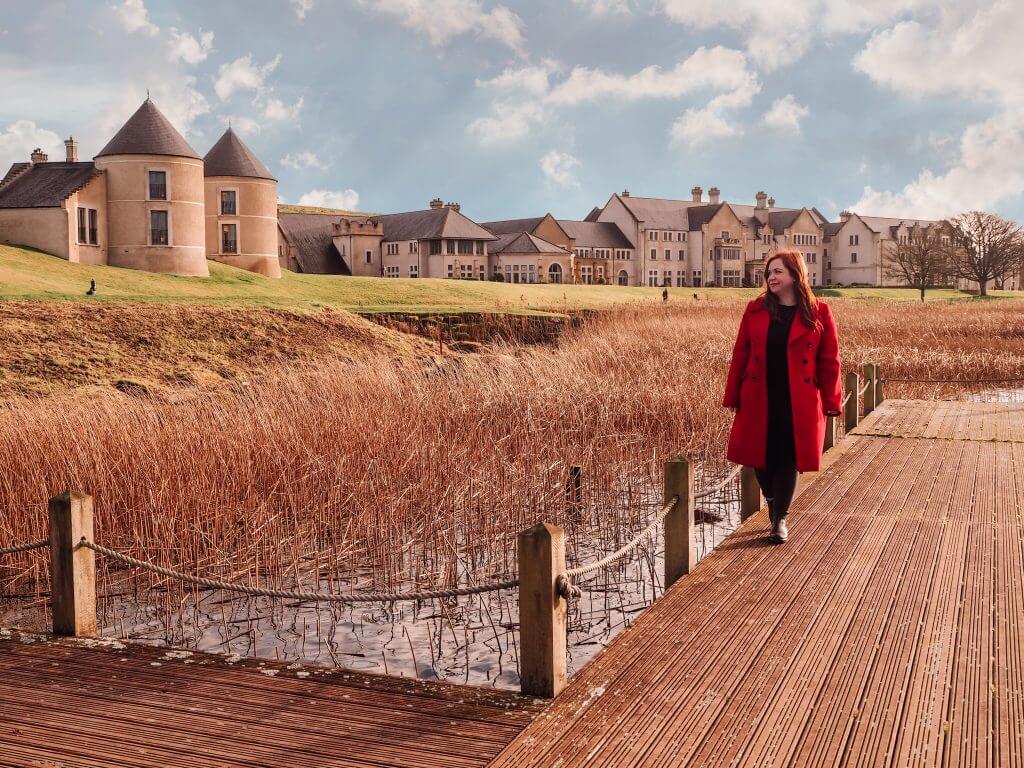 Travel blogger All about RosaLilla walking beside the lakeshore on a wooden jetty wearing a red coat. Hotel building can be seen in the background.
