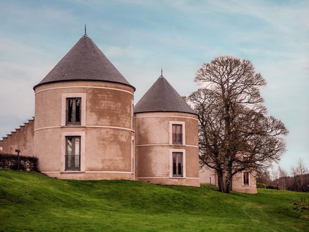 Circular buildings with turret style roofs standing beside a tree on green grass.