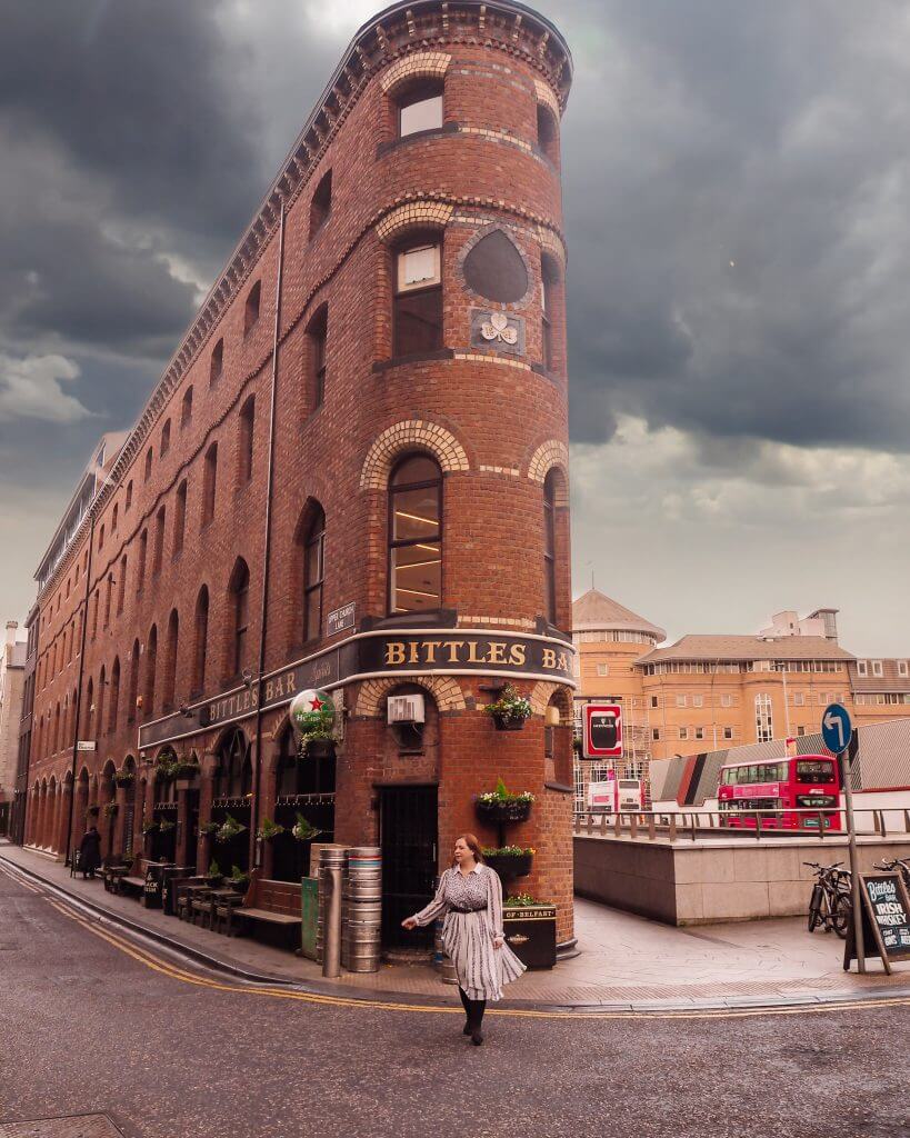 Woman in a white dress standing in front of Bittles Bar in Belfast as a red bus drives by.