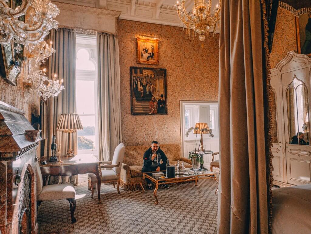 A man sits on a patterned, fringed sofa in a grand, classically decorated room at Ashford Castle, holding a glass of champagne. He is centered in the frame, positioned behind a glass-topped coffee table that holds a bottle in a silver chiller and a tall white orchid.
The lavish suite is filled with historical details, including ornate peach and cream damask wallpaper, a large crystal chandelier, and several framed paintings on the walls. To the left, a reddish-brown marble fireplace and a dark wood writing desk with a shaded lamp are visible. On the right, the scene is framed by the heavy, neutral-toned drapes of a large canopy bed and a white arched wardrobe.