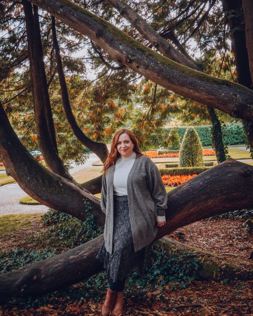 A woman with long auburn hair leans against a thick, mossy tree trunk that grows at a sharp horizontal angle. She is smiling at the camera, dressed in a white turtleneck, a grey cardigan, and a long dark patterned skirt.
The background features a formal garden with manicured hedges, a gravel path, and vibrant orange flowerbeds. Large, overhanging evergreen branches frame the shot, creating a natural canopy over the forest floor covered in fallen leaves and ivy.