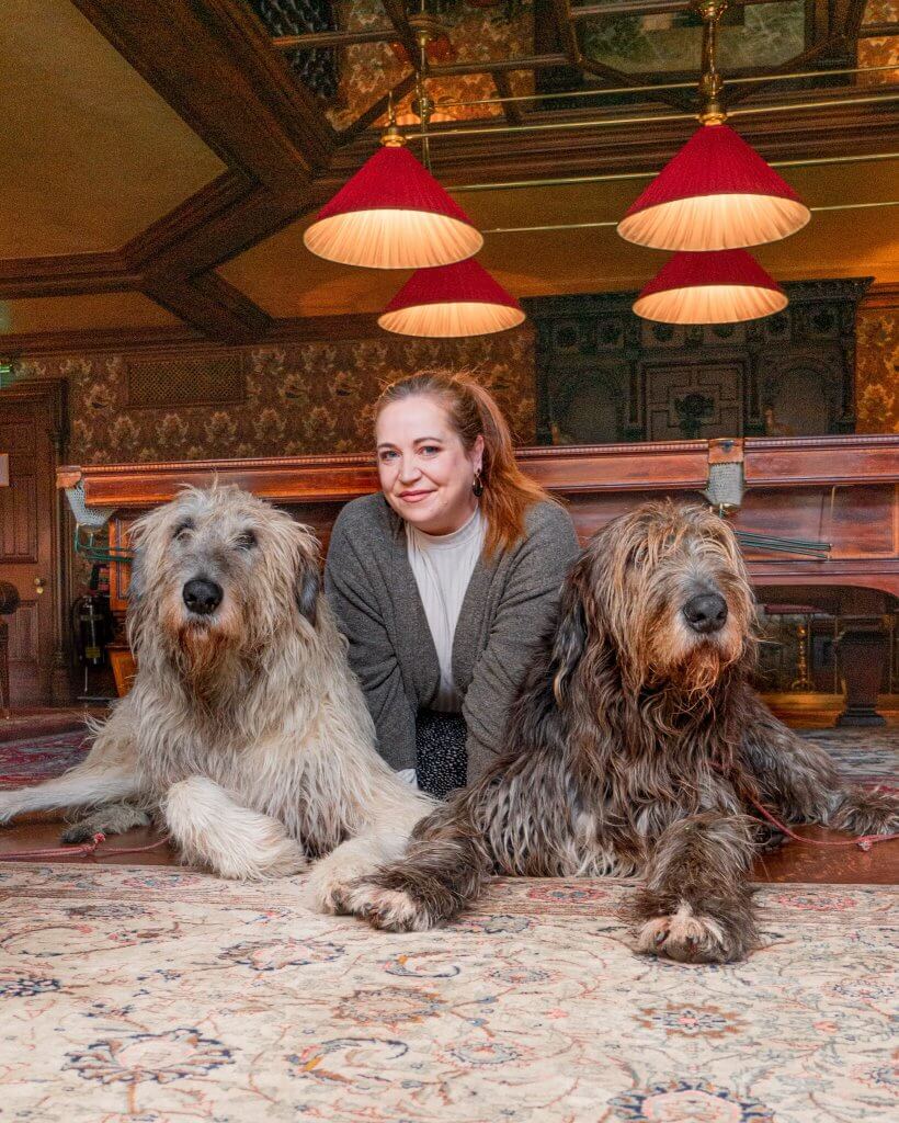A woman with long auburn hair sits on a patterned rug between two large, shaggy Irish Wolfhounds inside a grand, wood-paneled room at Ashford Castle. One dog has a light grey coat, while the other is a darker brindled brown; both are lying down and looking toward the camera.
Behind them, a large wooden billiards table sits under four vibrant red, cone-shaped pendant lights. The room is decorated with ornate floral wallpaper and dark wood accents, creating a warm, historic atmosphere.