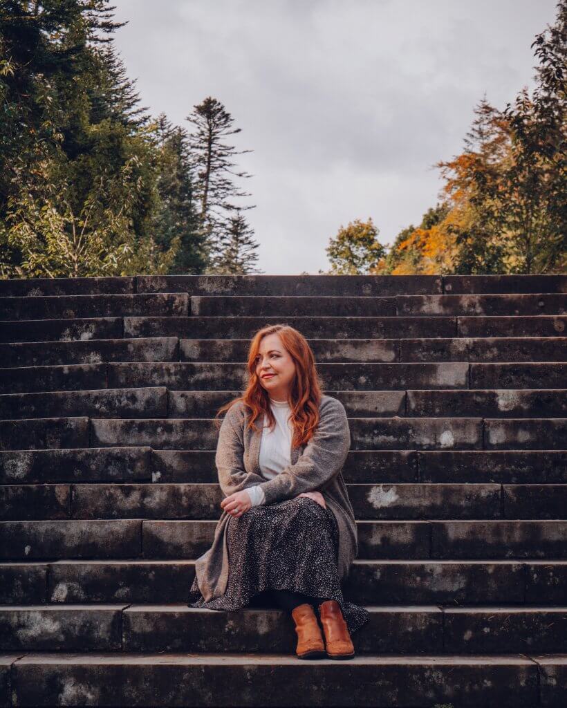 A woman with long auburn hair sits in the center of a wide, grand stone staircase outdoors. She is wearing a white turtleneck, a long grey cardigan, a dark patterned midi skirt, and tan leather boots.
She is looking away from the camera toward the left with a soft smile. Behind her, the staircase ascends toward a lush backdrop of tall evergreen and deciduous trees with some yellowing autumn leaves under an overcast sky. The weathered stone steps fill the lower and middle portions of the frame, emphasizing the scale of the structure on the grounds of Ashford Castle.