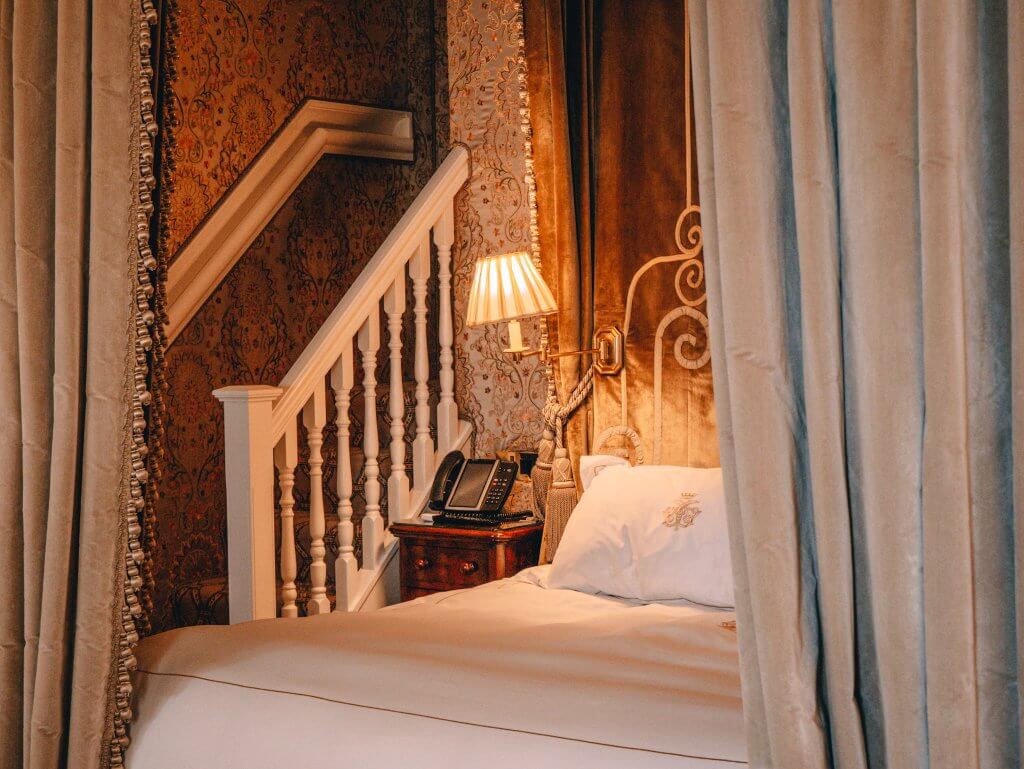 A close-up view of a grand canopy bed in a luxurious suite at Ashford Castle. The bed features crisp white linens with an embroidered crest and a tall, golden-brown velvet headboard with intricate scrollwork.
Heavy, neutral-toned drapes with decorative fringe frame the bed. To the left, a small dark wood nightstand holds a classic shaded lamp and a black telephone. In the background, a white wooden staircase with a banister leads upward against a wall covered in ornate peach and cream damask wallpaper.