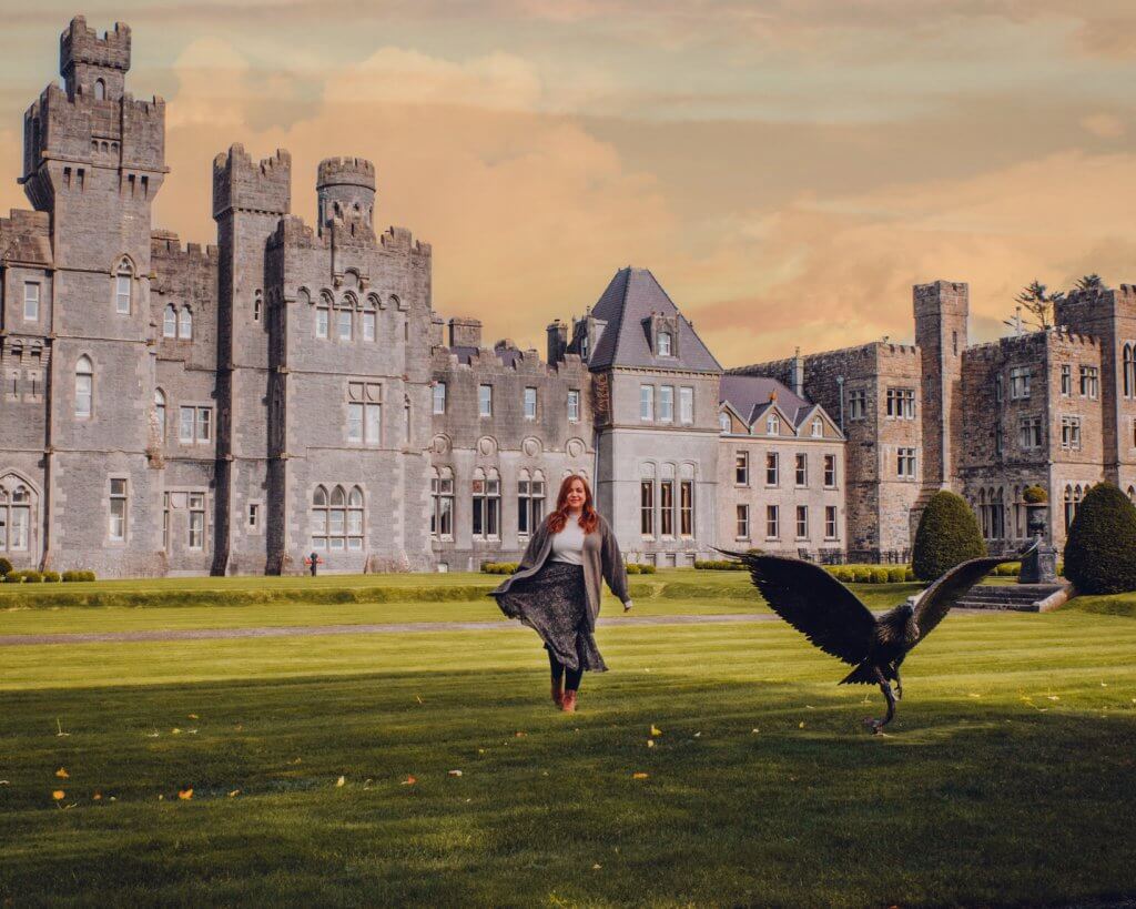 A woman with long auburn hair walks across a lush green lawn in front of a sprawling, Ashford Castle under a warm, golden-hour sky. To her right, a large bird of prey with dark feathers is captured mid-flight, its wings fully extended as it flies low over the grass. The woman is wearing a light-colored top, a long dark patterned skirt, and a grey cardigan that flows as she moves. The castle features numerous grey stone towers, battlements, and arched windows, creating a majestic and historic atmosphere.
