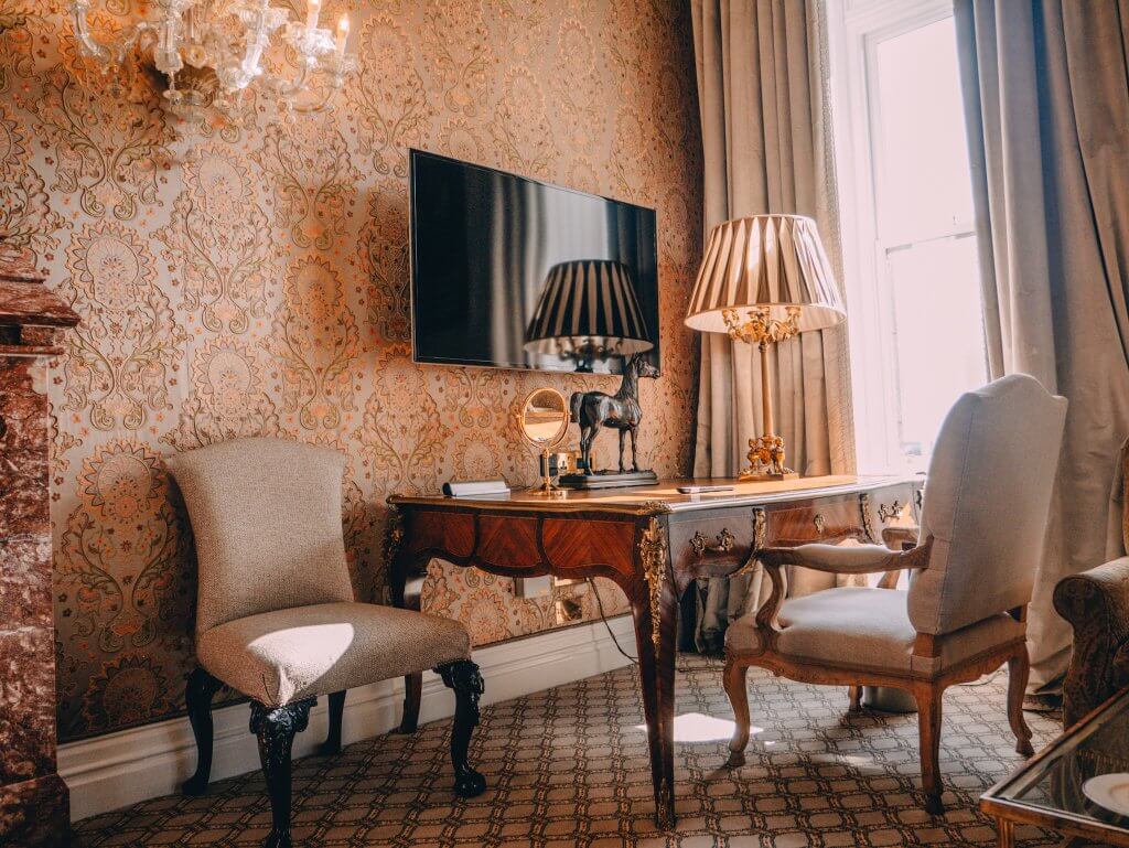 A polished dark wood writing desk with ornate gold accents sits against a wall covered in intricate peach and cream damask wallpaper in a room at Ashford Castle. On the desk, there is a classic shaded lamp with a gold base, a small bronze horse statue, and a round gold vanity mirror.
A flat-screen television is mounted on the wall above the desk, reflecting a nearby window. To the left, a neutral upholstered chair with dark, carved wood legs stands near a reddish marble fireplace. To the right, another identical chair is tucked into the desk next to a tall window draped with heavy tan curtains.