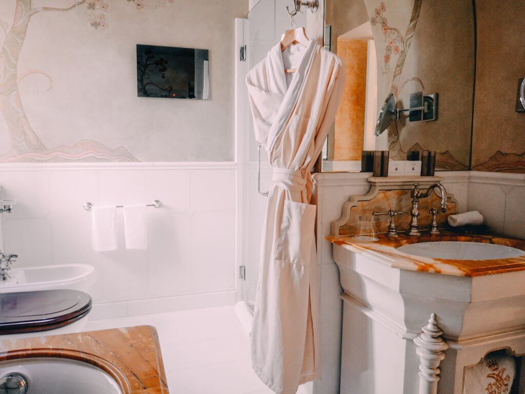 Bathroom at Ashford Castle Hotel. An elegant, marble-topped vanity stands in a bright, luxurious bathroom at Ashford Castle. A plush, cream-colored bathrobe hangs from a hook on a glass shower door in the center of the frame.
The vanity features ornate silver faucets and is set against a wall with delicate, hand-painted floral murals. To the left, a white tiled wall holds a silver towel rack with two folded white towels, and a glimpse of a dark-lidded commode is visible in the foreground. The room is filled with soft, natural light, highlighting the classical and refined decor.