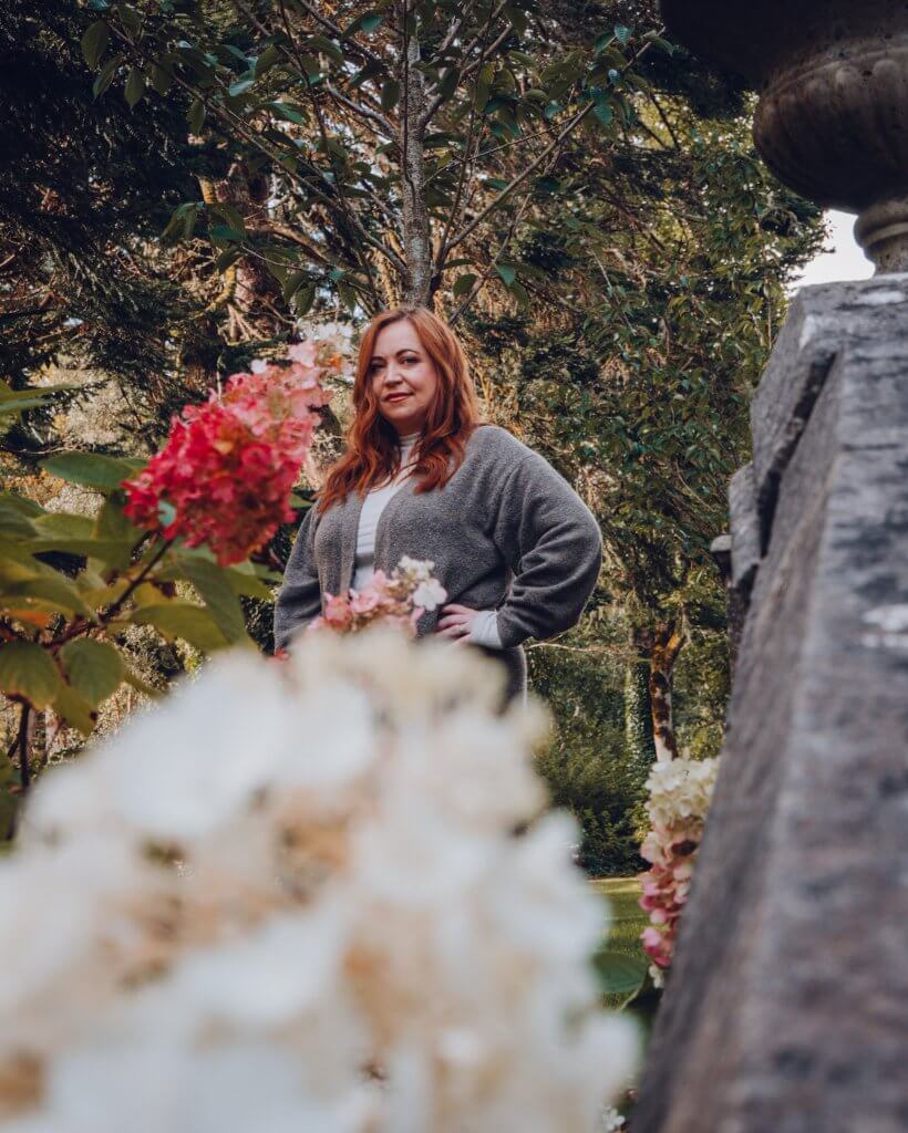 A woman with long auburn hair stands in a lush garden, viewed through a soft-focus foreground of pink and white hydrangea blossoms. She is wearing a white turtleneck and a long grey cardigan, with one hand resting on her hip as she looks toward the camera.
The background is filled with a variety of green trees and dappled sunlight, creating a serene, natural atmosphere on the grounds of Ashford Castle. A stone structure, likely part of a garden staircase or wall, is partially visible on the right side of the frame.