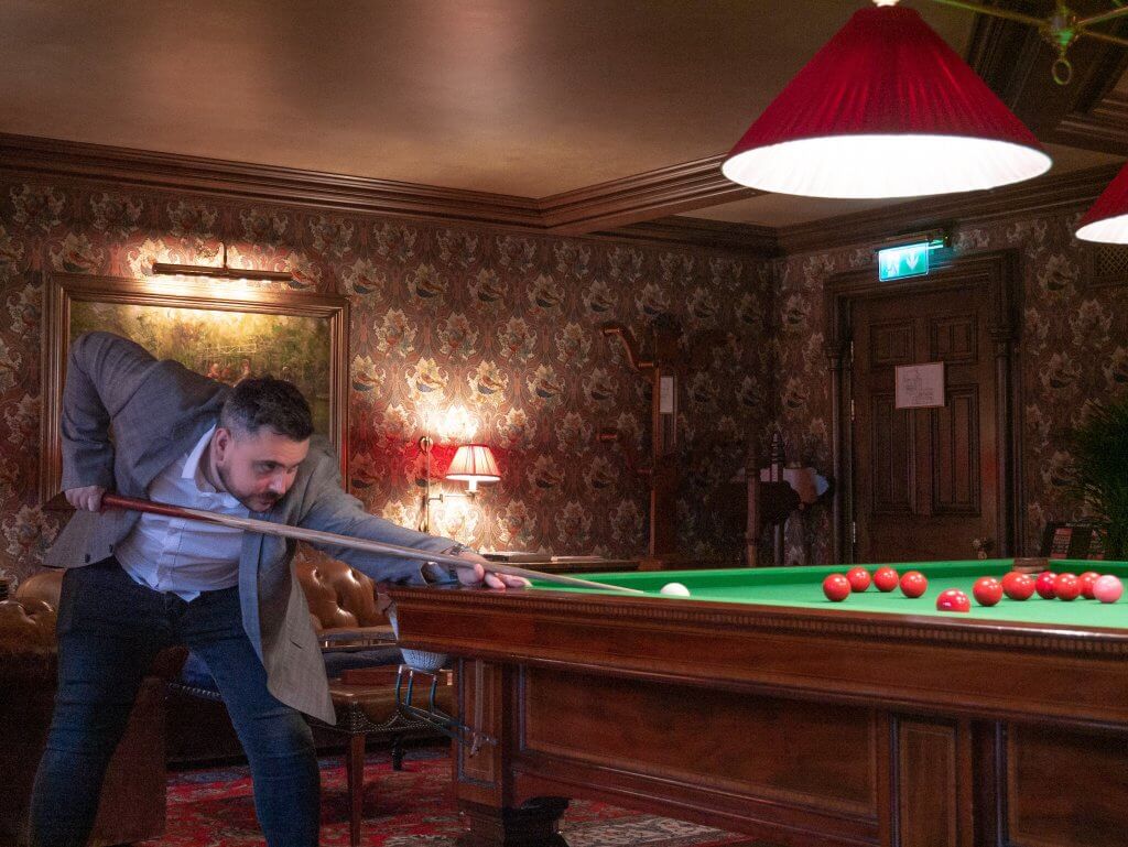 A man in a grey blazer and dark jeans leans over a large snooker table, concentrating as he prepares to take a shot with a wooden cue. He is aiming at a white cue ball on a green felt surface, with several red balls clustered nearby.
The room, located at Ashford Castle, features a luxurious and traditional aesthetic with intricate floral wallpaper, dark wood paneling, and a tufted leather sofa in the background. A large, red cone-shaped lamp hangs directly over the table, casting a focused glow on the game.