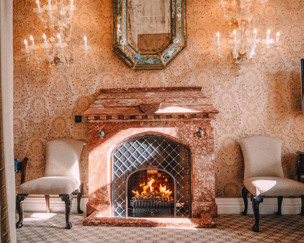 Open roaring fire in a bedroom at Ashford Castle. A close-up of an ornate, reddish-brown marble fireplace with a warm fire burning in the grate. The fireplace is set against a wall featuring intricate, light-colored damask wallpaper and is flanked by two elegant upholstered chairs with dark wood legs. Above the mantel, a decorative octagonal mirror is mounted between two glowing crystal wall sconces, all located within a luxurious room at Ashford Castle.