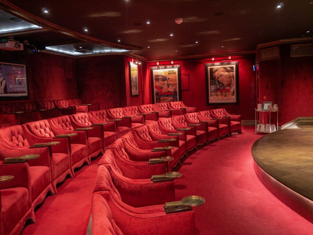 Luxury travel Ireland, cinema room at Ashford Castle Ireland. A wide shot of a luxurious, private cinema at Ashford Castle, featuring several curved rows of plush, red velvet armchairs. Each armchair has a tufted back and a small, built-in brass tray on the armrest.
The room is decorated with deep red velvet walls and a matching red carpet. Several framed classic movie posters, including Chisum and Dirty Harry, are mounted on the walls and illuminated by overhead spotlights. To the right, a curved dark wood stage area is visible, while a small service cart stands in the background.