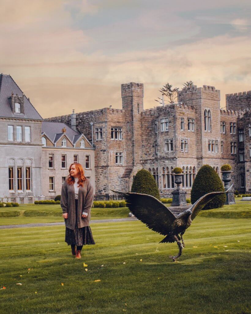 A woman with long auburn hair walks across a lush green lawn at Ashford Castle, looking toward a large bronze sculpture of an eagle with its wings fully spread. She is dressed in a white turtleneck, a long grey cardigan, a dark patterned midi skirt, and tan boots.
The grand, historic stone castle serves as the backdrop, its various wings and turrets illuminated by the soft, warm glow of a late afternoon sky. Manicured hedges and decorative stone planters are visible in the distance, completing the serene and majestic landscape.