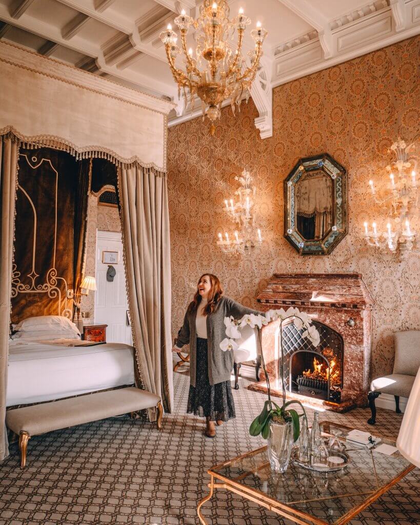 A woman with long auburn hair stands in a lavish, high-ceilinged bedroom at Ashford Castle, looking up in awe with a wide smile. She is wearing a white turtleneck, a long grey cardigan, and a dark patterned midi skirt.
To her left is a grand canopy bed with tall, draped curtains and an ornate dark velvet headboard. On the right, a marble fireplace has a warm fire burning inside, topped by a decorative mirror and flanked by glowing crystal wall sconces. A large crystal chandelier hangs from the white coffered ceiling. The room is decorated with intricate floral wallpaper and a patterned carpet, featuring a glass coffee table with white orchids in the foreground.