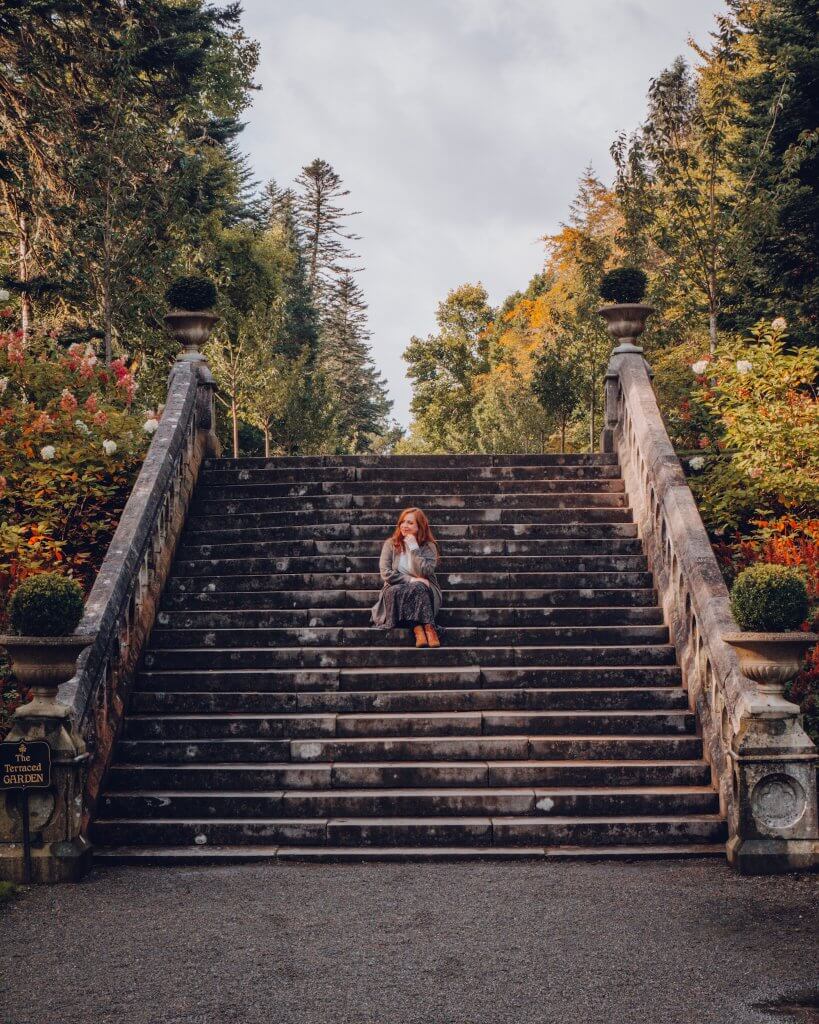 A woman with long auburn hair sits thoughtfully on a wide, weathered stone staircase leading up into a lush forest. She is wearing a grey cardigan, a dark patterned skirt, and tan boots, resting her chin on her hand while looking off to the side.
The grand staircase is flanked by stone banisters topped with decorative urns containing manicured shrubs. Vibrant autumn foliage and tall evergreen trees frame the scene under a soft, overcast sky. A small sign at the bottom left identifies the location as "The Terraced Garden," a feature of the grounds at Ashford Castle.