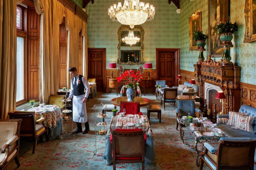 Afternoon tea in the Connacht Room at Ashford Castle. A server in a white apron and black vest prepares a tiered stand of pastries at a small table in a grand, sunlit tea room at Ashford Castle. The room is filled with elegant, historic details, including high ceilings with a large crystal chandelier, seafoam-green patterned wallpaper, and a dark wood-carved fireplace.
The space is arranged with several intimate seating areas featuring ornate blue and red velvet chairs, patterned sofas, and tables set for afternoon tea. Tall windows with heavy cream-colored drapes line the left wall, while large gold-framed portraits and a decorative mirror hang on the far walls. An ornate blue and white vase with vibrant red flowers serves as a centerpiece on a round table in the middle of the room.