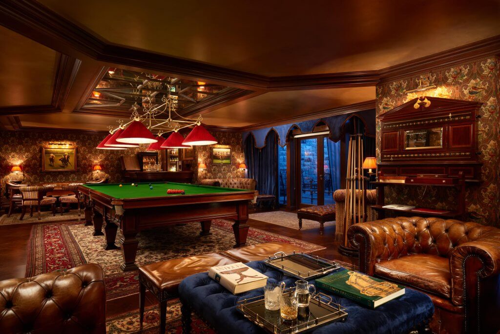 A luxurious billiard room at Ashford Castle featuring a large, professional snooker table with a green baize top and red balls arranged at one end. A row of red cone-shaped lamps hangs from a mirrored ceiling panel, casting a warm glow over the table.
The room is decorated with ornate floral wallpaper, dark wood paneling, and several tufted brown leather armchairs. In the foreground, a blue velvet ottoman holds books and a tray with crystal glasses. Rich red patterned rugs cover the dark wood floor, and large blue drapes frame a glass door leading to an outdoor stone patio in the background.