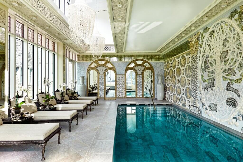 The spa and pool at Ashford Castle Hotel the ultimate in luxury travel ireland. A brightly lit, opulent indoor pool at Ashford Castle features a long, rectangular swimming pool with turquoise water. Along the left side, four plush, cream-colored lounge chairs are arranged, each accompanied by a small side table with white orchids.
The far wall is adorned with a massive, intricate mosaic depicting a "Tree of Life" filled with various animals, including birds and a hare. Two arched mirrors at the end of the room reflect the bright, airy space. The ceiling is highly decorative with intricate patterns and features two large, ethereal beaded chandeliers hanging over the pool area. High windows on the left allow natural light to fill the room, highlighting the detailed tile work and serene atmosphere.