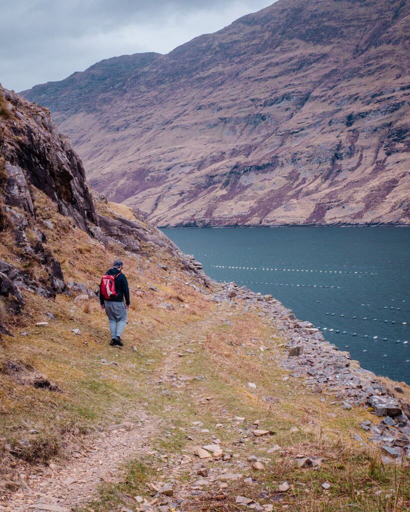 Man in a grey tracksuit and red backpack walking along a rocky mountain path on the Killary harbour coastal walk on a connemara hike
