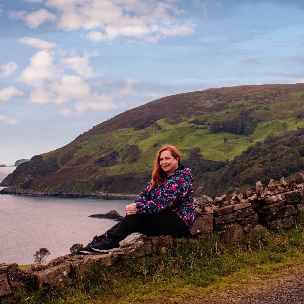 Woman sitting on a stone wall overlooking dramatic scenery at Murlough Bay known as Slavers Bay in Game of Thrones