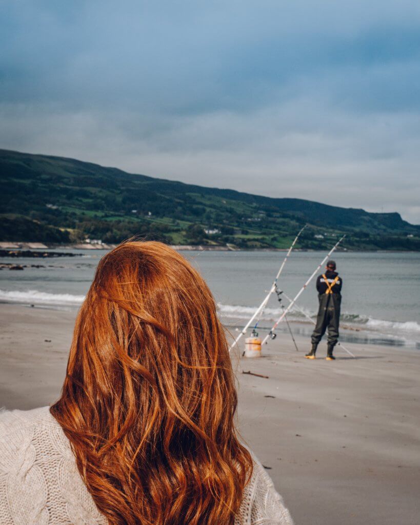 Woman with red hair watching a man fishing on the beach at Ballygally in County Antrim Northern Ireland