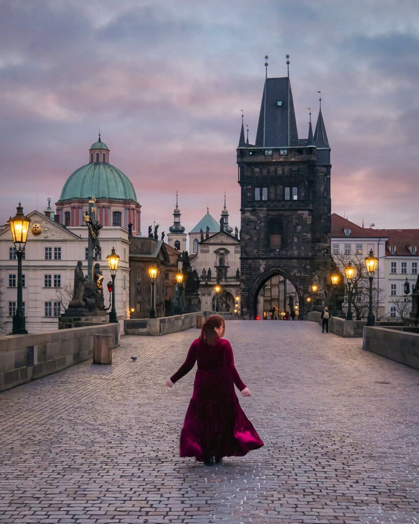 Woman in a ;ong red velvet dress walking along Charles Bridge at sunrise