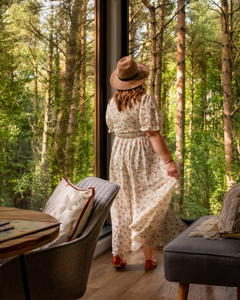 Nicola Lavin, Irish travel blogger with Our Unique Stays, wears a floral dress and looks out the window of Burrenmore Nest treehouse.