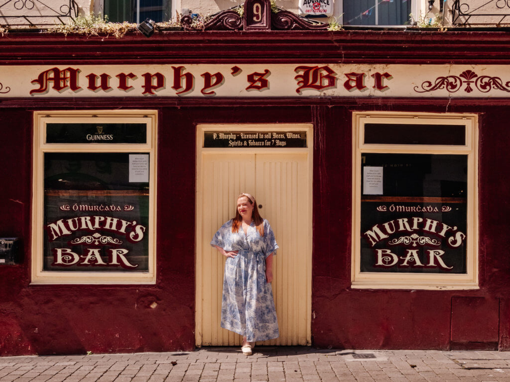 A woman in a blue floral dress stands in front of Murphy's Bar, one of the oldest pubs in Galway.