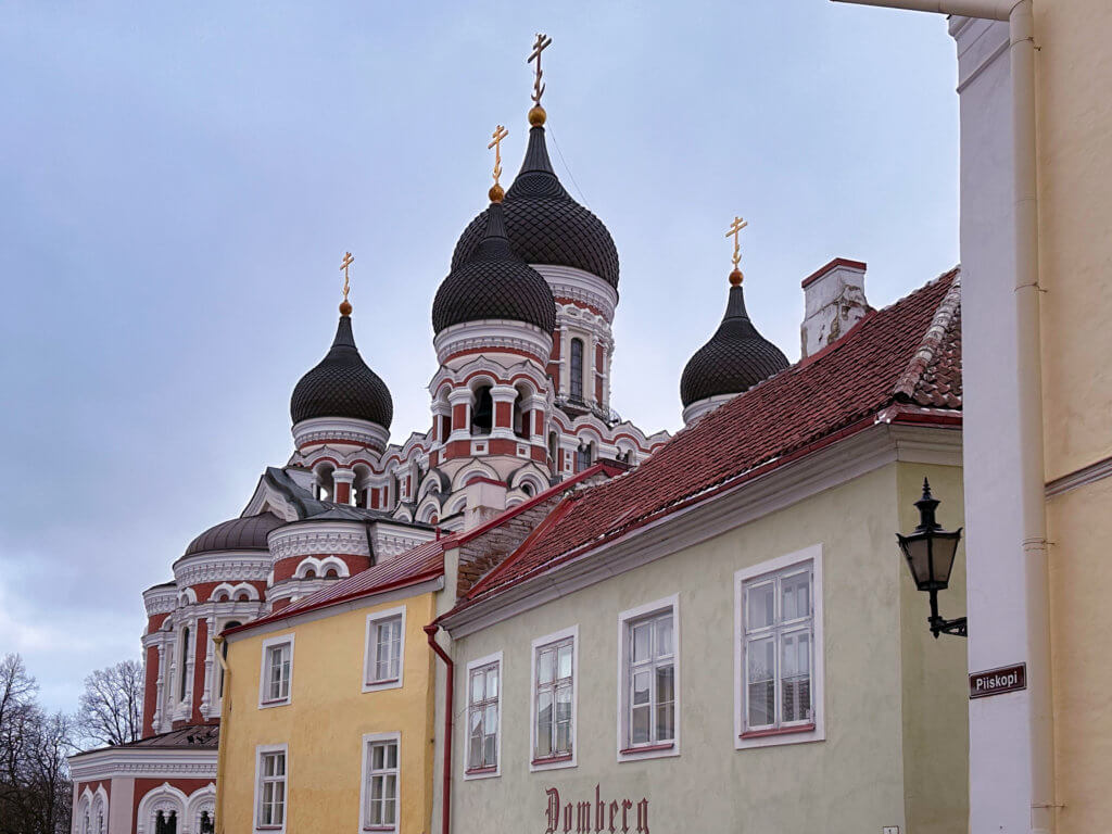 An eye-level, landscape-oriented photograph showing the black onion domes of the Alexander Nevsky Cathedral in Tallinn, Estonia, rising above a row of colorful historic buildings.
In the foreground, a light green building with white-framed windows features the word "Domberg" in dark red lettering near the bottom. Next to it is a yellow building, also with white windows and a red-tiled roof. A white building with a brown "Piiskopi" street sign on the corner frames the right side of the shot. The cathedral towers behind these structures, showcasing its red brick and white stone facade and multiple domes topped with golden crosses against an overcast, pale blue sky.