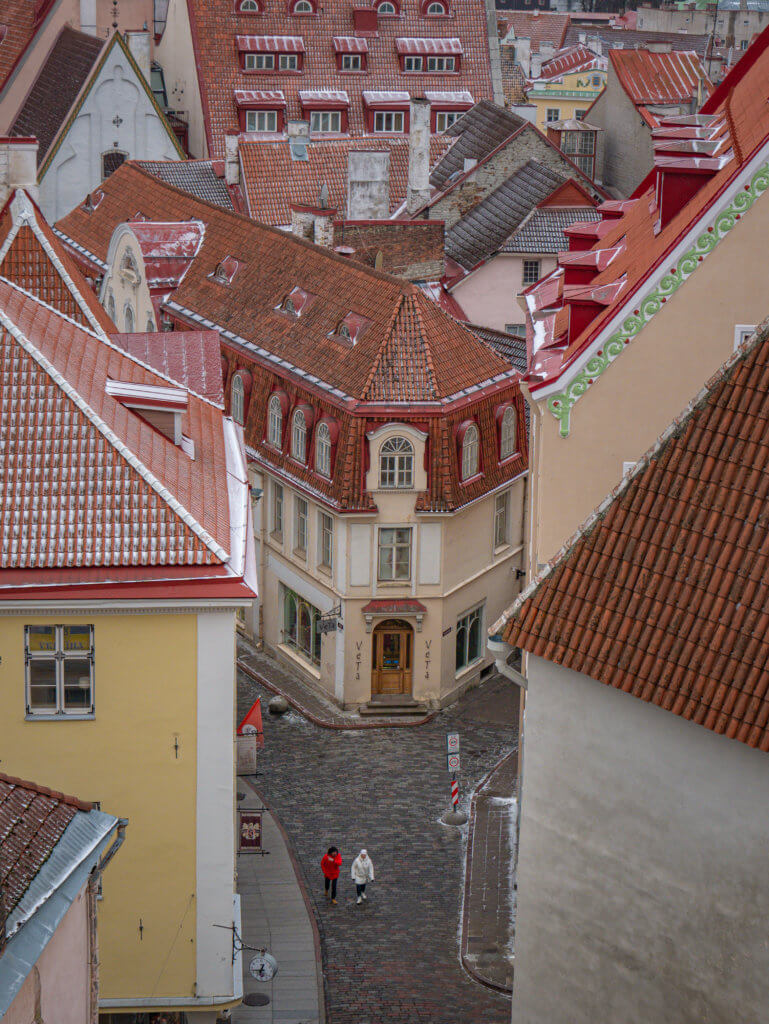Rooftop views in Old Town Tallinn Estonia. An elevated, high-angle shot captures a narrow cobblestone street in a European Old Town, likely Tallinn, Estonia, during winter.
At the center, a triangular, cream-colored building with a red gambrel roof and decorative dormer windows sits at a fork in the road. Its ground floor features a wooden door with "Veta" signage. To the left, a bright yellow building anchors the foreground, while the right is framed by a tall, textured white wall.
Two people, one in a bright red jacket and the other in white, walk along the cobblestones below. The background is a dense, rhythmic pattern of terracotta-tiled rooftops dusted with light snow, interspersed with chimneys and various window styles, creating a cozy, medieval atmosphere.