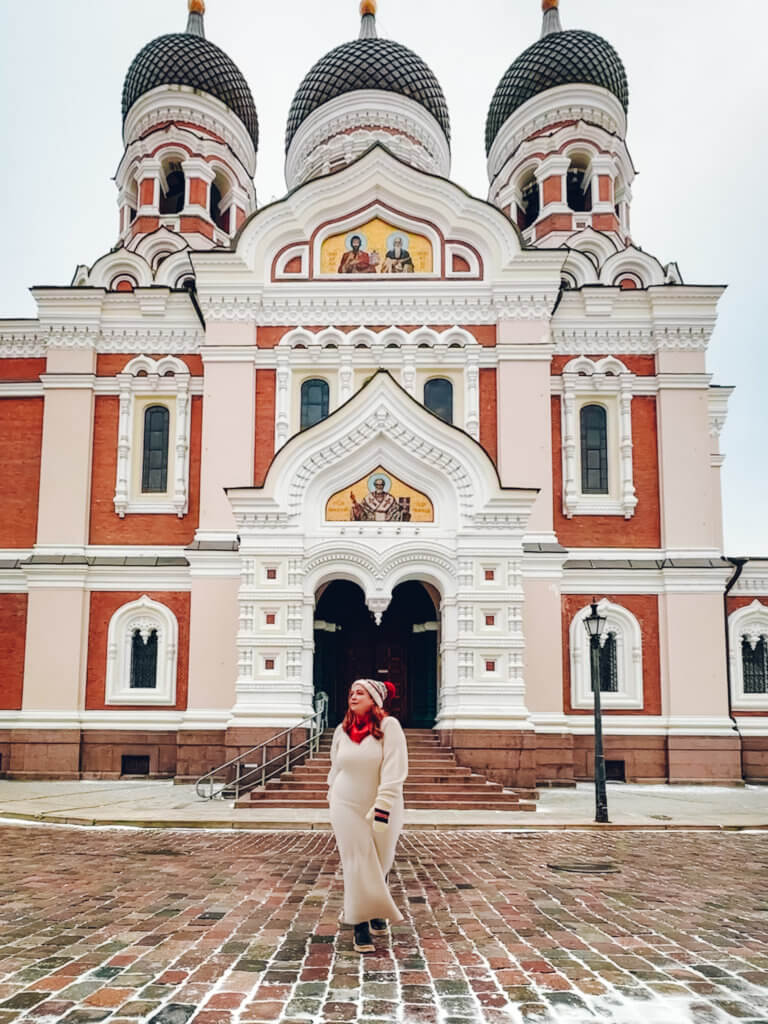 A portrait-oriented photo shows a woman in a long white dress and a red-and-white knit hat standing in front of the Alexander Nevsky Cathedral in Tallinn, Estonia. The cathedral features a red and white facade with elaborate white trim, arched windows, and three large black onion domes topped with golden crosses. Golden religious icons are visible in mosaics above the arched entrance and on the upper gables. The woman is standing on a snow-dusted cobblestone square, looking slightly to the side. The sky above is overcast and flat grey.