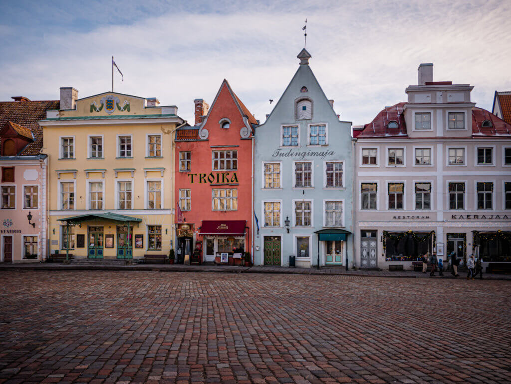 Town Square in Old Town Tallinn in winter. A wide-angle shot of a historic cobblestone town square in Tallinn, Estonia, featuring a row of four colorful, medieval-style buildings. From left to right: a pale yellow building with a green awning, a narrow terracotta-red building with "TROIKA" written in gold letters, a light blue gabled building labeled "Tudengimaja," and a white building with red roofing housing the "Kaerajaan" restaurant. The square is mostly empty under a soft, overcast sky, with a few people walking in the distance on the far right.