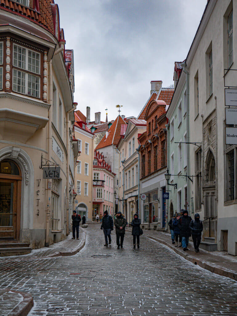 A portrait-oriented photo shows a narrow, cobblestone street in Tallinn, Estonia, during winter. The street is wet and dusted with light snow. Several people in dark winter coats walk away from the camera toward the center of the frame.
The street is lined with historic, multi-story buildings in various colors, including cream, pale yellow, and light green. On the left, a building features a prominent arched doorway and a sign for "VeTa." In the background, steep red-tiled roofs with small dormer windows and decorative weather vanes rise against a flat, overcast grey sky.