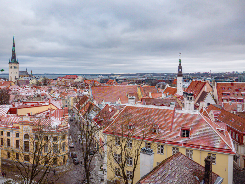 A wide, high-angle panoramic view overlooks the historic Old Town of Tallinn, Estonia, under a heavy, overcast gray sky.
The foreground and midground are filled with a dense cluster of medieval-style buildings featuring steep, orange-terracotta tiled roofs lightly dusted with snow. To the left, a prominent cream-colored building with ornate white trim sits at a street corner where several cars are parked. Bare, dark-branched trees rise from the street level, reaching toward the rooftops.
The skyline is punctuated by two notable landmarks:
On the far left, the tall, slender, dark green spire of St. Olaf’s Church rises above a white stone tower.
To the right, the white tower of the Town Hall is visible with its distinctive black-capped spire.
In the far distance, the Baltic Sea is visible as a thin blue-gray line on the horizon, with the faint silhouettes of modern port cranes and ships.