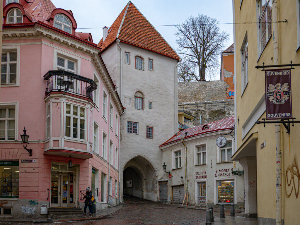 Pikk Jalg cobblestone street in Tallinn during winter. A ground-level, wide-angle photograph of a historic street intersection in Tallinn, Estonia. The scene is dominated by a tall, white medieval gatehouse building with a steep, orange-tiled pyramidal roof and a large pointed archway at its base. To the left stands a three-story pastel pink building with a decorative white bay window and a black wrought-iron balcony.
The cobblestone street is damp and slopes gently upward toward the archway. On the right, a smaller two-story white building features signs for "VALUUTA VAHETUS" and "MONEY CHANGE," topped by a round exterior clock. In the far background, a bare winter tree rises above a stone defensive wall. The sky is a flat, overcast grey.