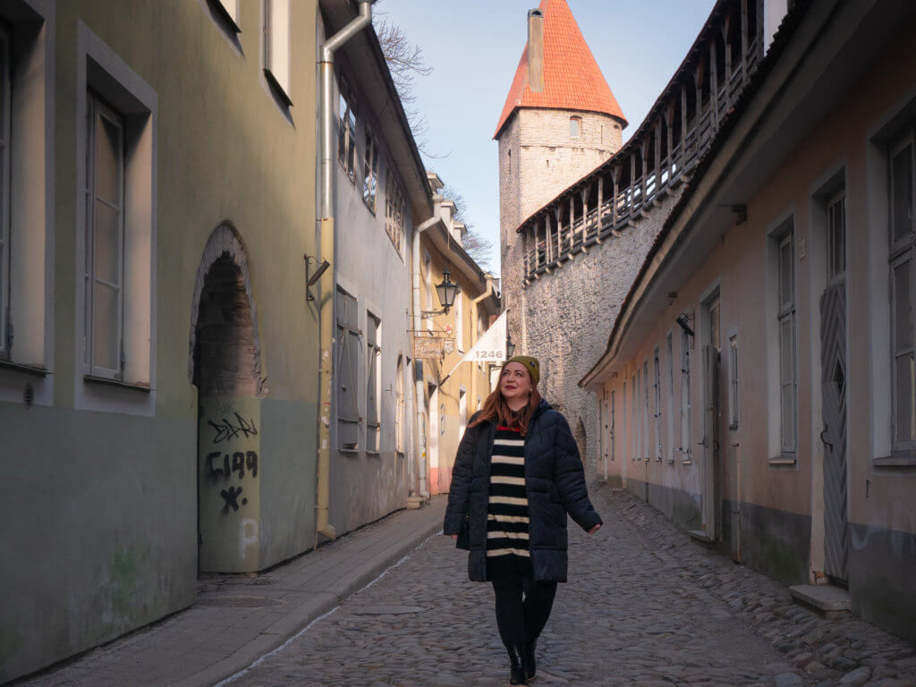 A portrait-oriented photo shows a woman walking toward the camera on a narrow, cobblestone street in Tallinn, Estonia. She is wearing a dark puffer coat over a black-and-white striped dress and a mustard-yellow beanie.
On the right side of the frame, a high medieval stone wall stretches into the distance, topped with a covered wooden defensive gallery. A tall stone tower with a peaked red-tiled roof rises at the end of the wall. To the left, the street is lined with historic buildings in shades of muted green and grey. The sky is a soft, pale blue, and the scene is illuminated by gentle, natural light.