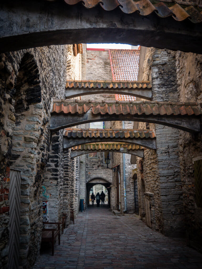 A portrait-oriented photo looking down St. Catherine's Passage, a narrow medieval alleyway in Tallinn, Estonia. The path is paved with uneven reddish bricks and stones, flanked by ancient, high walls made of rough-hewn grey limestone.
The alley is famous for its series of large stone buttress arches topped with small, orange-tiled roofs that span across the passage at various intervals. In the distance, through a low stone archway at the end of the alley, the silhouettes of three people are visible walking toward the light. On the left wall, a couple of dark wooden benches sit in the shadows.