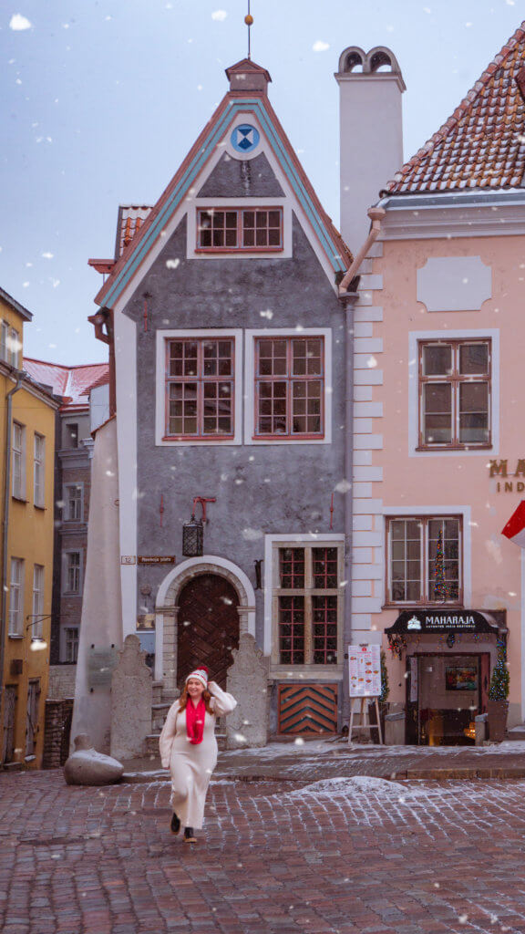 A portrait-oriented photo of a woman walking across a damp cobblestone square in Tallinn, Estonia, while light snow falls. The woman is smiling and wearing a long white sweater dress, a bright red scarf, and a red and white knit hat.
Behind her stands a narrow, grey medieval house with a steep gabled roof, a dark arched wooden door, and a street sign that reads "Raekoja plats." To the right is a light pink building housing the "Maharaja" Indian restaurant. The ground is partially covered with thin patches of slush and snow.