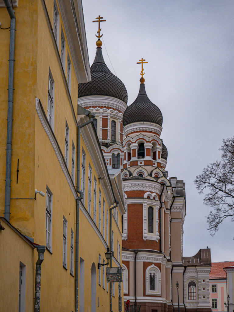 A portrait-oriented photo taken from a low angle on a street in Tallinn, Estonia, looking up at the Alexander Nevsky Cathedral. On the left, a tall, pale yellow building with white-framed windows and grey downspouts recedes into the distance.
The cathedral rises behind it, featuring its characteristic red brick and white stone facade. Two of its large, dark grey onion domes are visible, each topped with an ornate golden cross. The sky is a flat, overcast grey, highlighting the intricate architectural details and vibrant colors of the cathedral’s upper tiers.