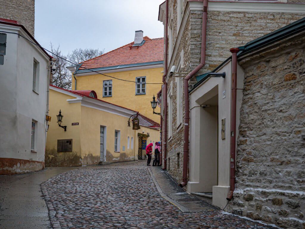Medieval streets in Old Town Tallinn Estonia. ground-level view of a winding cobblestone street in Tallinn, Estonia, on an overcast day. The street curves uphill toward the center of the frame, bordered by historic stone and plastered buildings. On the right, a textured stone wall with several copper-colored downspouts and a white-framed doorway dominates the foreground. In the center, a bright yellow building with a red-tiled roof sits at the bend of the road, featuring dark-framed windows and a decorative wrought-iron street lamp. Two figures in brightly coloured winter jackets—one in pink and one in purple—stand near a doorway in the background. The cobblestones are uneven and appear slightly damp.