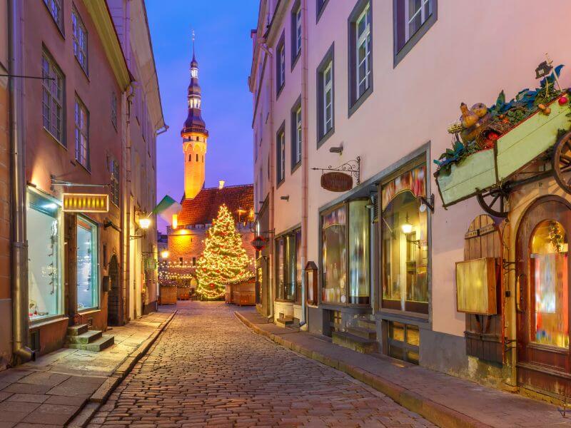 A narrow cobblestone street in Tallinn, Estonia, at dusk, leading toward a glowing Christmas tree in a town square. The street is lined with historic, multi-story buildings in pastel shades of pink and cream, featuring charming storefronts and warm interior lights. In the background, the illuminated spire of the Tallinn Town Hall rises into a deep blue twilight sky. The scene is festive and cosy, capturing the magical atmosphere of the Old Town during the holiday season.