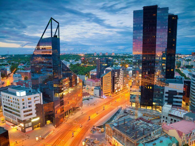 An elevated, high-angle evening shot of a modern city district in Tallinn, Estonia. Two prominent glass skyscrapers flank a wide, brightly lit main road with long exposure light trails from passing vehicles. The building on the left features a sharp, angular design with a triangular top, while the building on the right is a tall, dark rectangular monolith reflecting the warm orange glow of the city lights. In the background, the cityscape transitions into a dense green horizon under a cloudy, twilight blue sky.