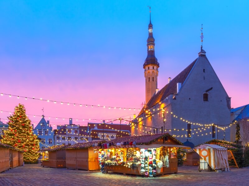 A wide shot of the Tallinn Christmas Market set against a vibrant pink and purple sunset. To the left, a tall, lit Christmas tree stands near several closed wooden stalls. In the center, one stall is brightly illuminated, displaying colorful winter hats and furs. To the right, the grand medieval Town Hall with its iconic slender spire rises into the darkening sky. Strings of warm globe lights crisscross overhead, connecting the tree and the historic buildings.