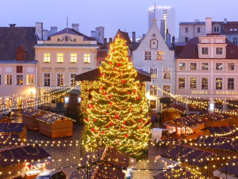 An elevated view of the Tallinn Christmas Market at dusk, featuring a large, brightly lit Christmas tree as the central focus.
The tree is covered in warm golden lights and red ornaments, with strings of fairy lights radiating outward like a canopy over rows of small wooden market stalls. The stalls have dark roofs also lined with lights. In the background, the colorful historic buildings of Town Hall Square are visible, with warm light glowing from their windows. The ground consists of wet cobblestones reflecting the festive lights, creating a cosy and magical winter atmosphere.