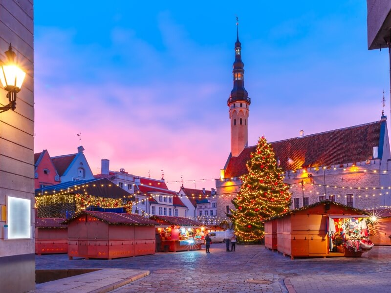 A vibrant evening view of the Tallinn Christmas Market in Estonia's Town Hall Square. The scene is set against a striking pink and blue twilight sky.
In the center, a large, glowing Christmas tree decorated with warm golden lights stands tall. Surrounding it are several wooden market stalls with red-trimmed roofs, some of which are open and displaying colorful goods. In the background, the historic Tallinn Town Hall, a medieval building with a distinct slender spire, is illuminated. String lights hang across the cobblestone square, and a few people are scattered throughout, adding to the festive, cosy atmosphere.