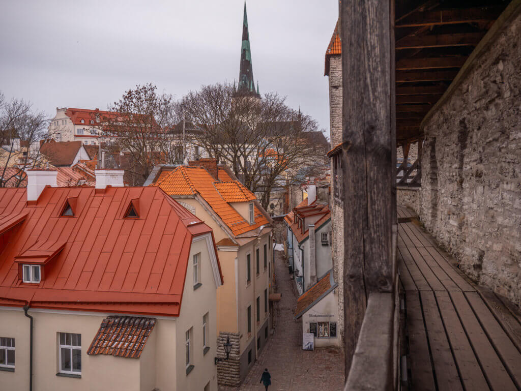 Wooden walkway of Hellemann Tower in Tallinn. A high-angle shot taken from a wooden medieval walkway looking down into a narrow cobblestone street in Tallinn, Estonia. On the left, several multi-story buildings with bright orange and red gabled roofs are huddled together, some featuring small triangular dormer windows. In the center, the street winds between the buildings, with a lone figure walking in the distance. The background is dominated by a dense cluster of historic rooftops and the tall, dark green spire of St. Olaf’s Church rising against a flat, overcast sky. The right side of the frame is bordered by the thick wooden support beams and stone wall of the elevated defensive gallery.