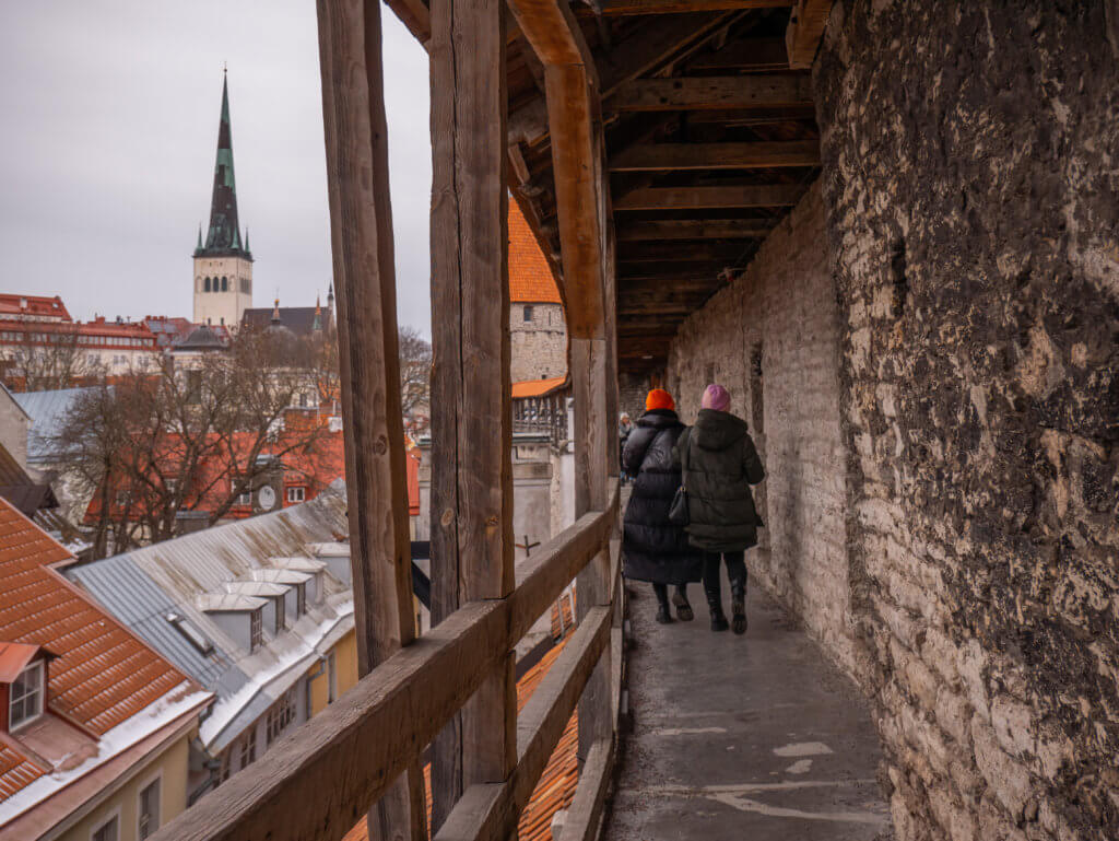 Wooden walking platform of Hellemann Tower in Tallinn in winter. A portrait-oriented photo taken from the perspective of an elevated medieval defensive walkway in Tallinn, Estonia. On the right, two people in dark winter coats and colorful hats (one orange, one pink) walk away from the camera along a narrow stone path. The path is protected on the right by a high, rough-hewn stone wall and on the left by a wooden railing and heavy vertical timber supports that hold up a peaked wooden roof.
The view to the left overlooks the snow-dusted red and grey rooftops of the Old Town. In the background, the tall, slender white tower and dark green spire of St. Olaf’s Church rise into a flat, overcast grey sky. A cylindrical stone defensive tower with a conical orange roof is visible further along the wall in the center of the frame.