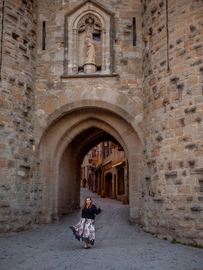A medium-angle shot captures a person wearing a dark top and a patterned, flowing skirt standing beneath a massive stone archway in Carcassonne, France.

The archway is built into a rugged, light-colored stone wall and features an ornate niche above the opening containing a religious statue. Through the arch, a narrow, rising cobblestone street is visible, lined with historic buildings. The person is positioned in the center of the walkway, looking toward the camera with a slight smile.